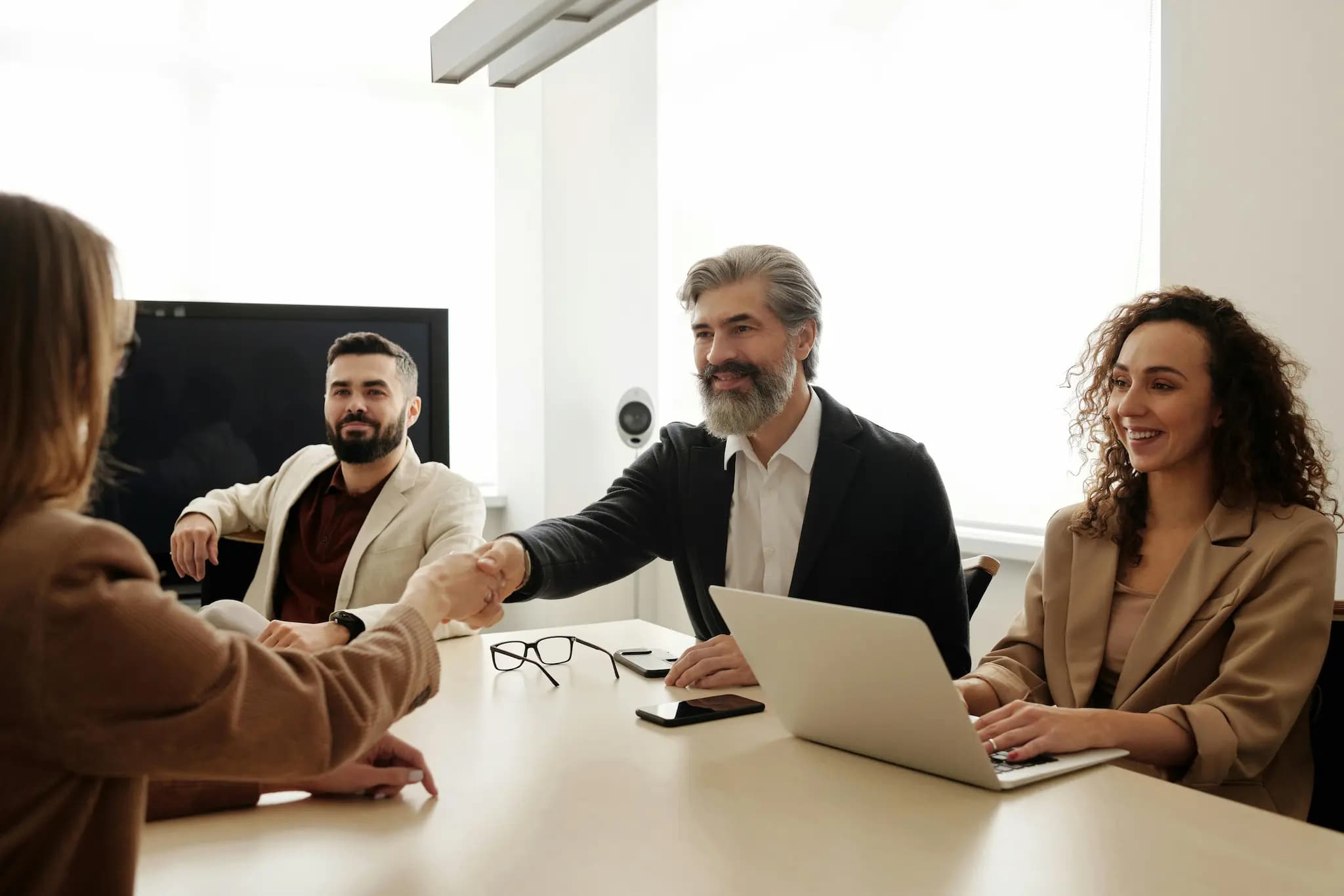 A person shakes hands across a conference table with three colleagues engaged in a business meeting, with laptops and a screen in view.